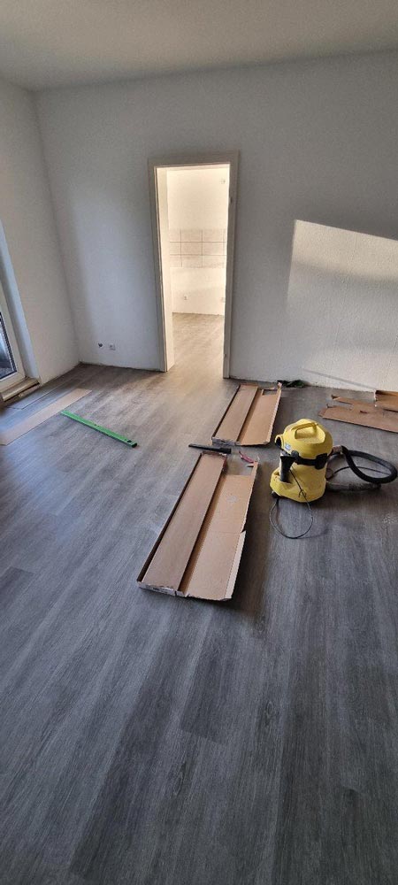 Ein leerer Raum mit neuem Laminatboden, gelber Staubsauger, Maßband und Kartonstücke am Boden.