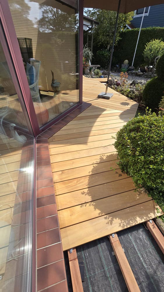 Holzterrasse an einem Eckfenster, umgeben von Sträuchern und einem Garten mit Steinen und Pflanzen.