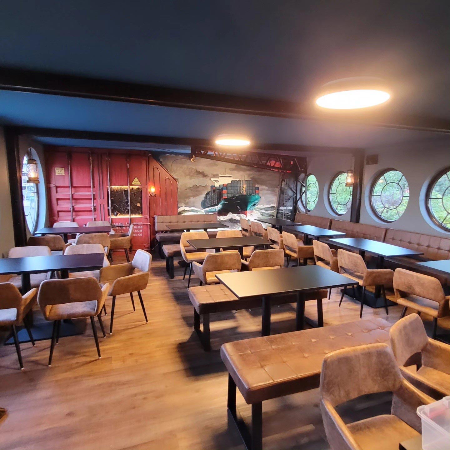 Gemütliches Café mit Holzboden, runden Fenstern und Wandmalerei eines Containerschiffs.
