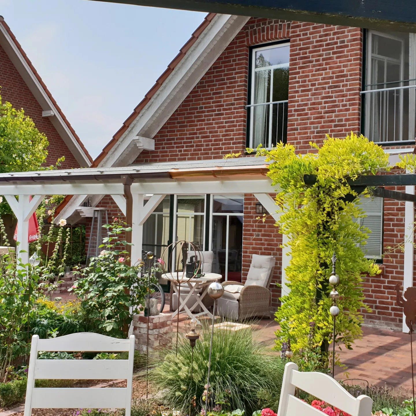 Gemütlicher Garten mit Terrasse, Backsteinhaus, viel Grün, weiße Möbel, Gelbes Rankwerk.