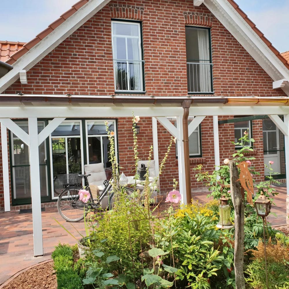 Zweistöckiges Backsteinhaus mit Garten, weißen Rahmen, Fahrrad und blühenden Blumen im Vordergrund.