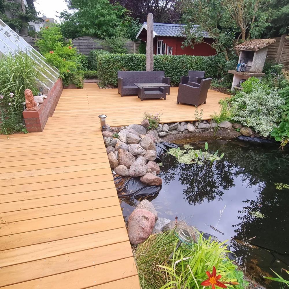 Holzterrasse mit Gartengarnitur, Teich, Felsen und üppiger grüner Bepflanzung im Hintergrund.