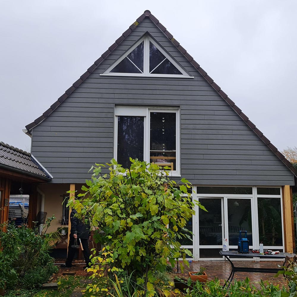 Graues A-Frame-Haus mit großen Fenstern, umgeben von Gartenpflanzen, unter bewölktem Himmel.