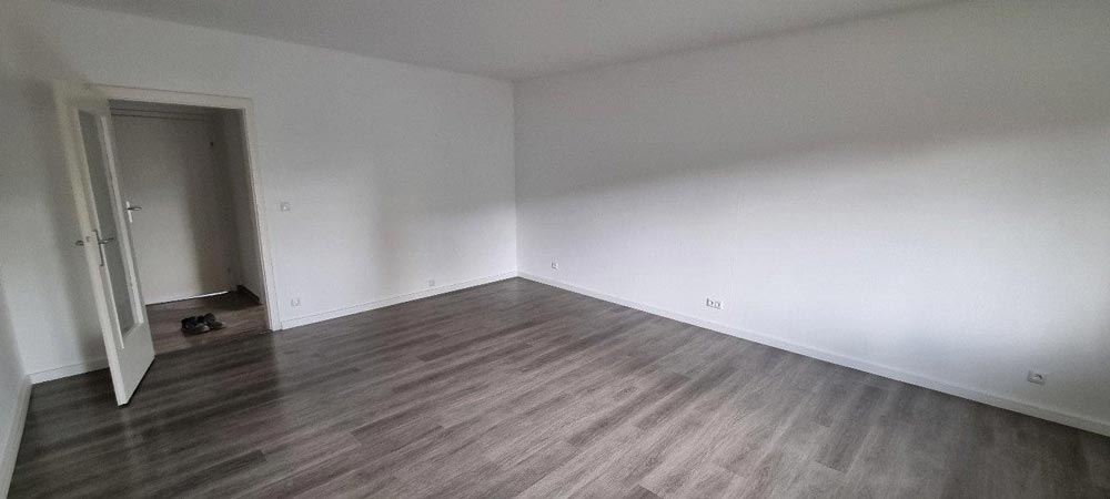 Leerer Raum mit grauem Laminatboden, weißen Wänden und einer offen stehenden Tür. Links sind Schuhe.