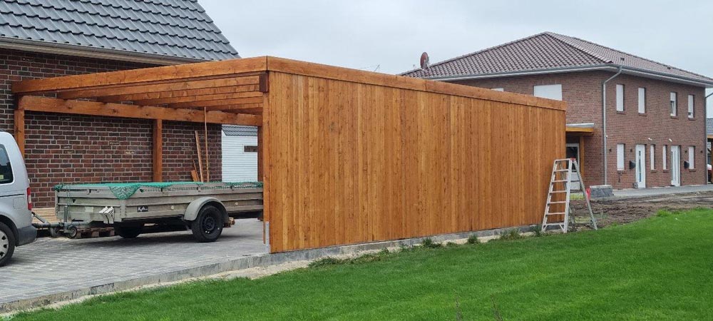 Holzcarport mit Anbau neben modernen Ziegelgebäuden, grauer Himmel, grüne Rasenfläche.