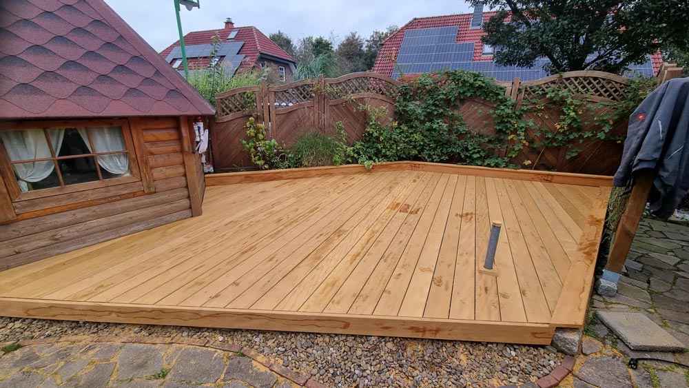 Holzterrasse mit gewinkeltem Design neben einem Holzhaus, im Hintergrund ein Garten mit Zaun.