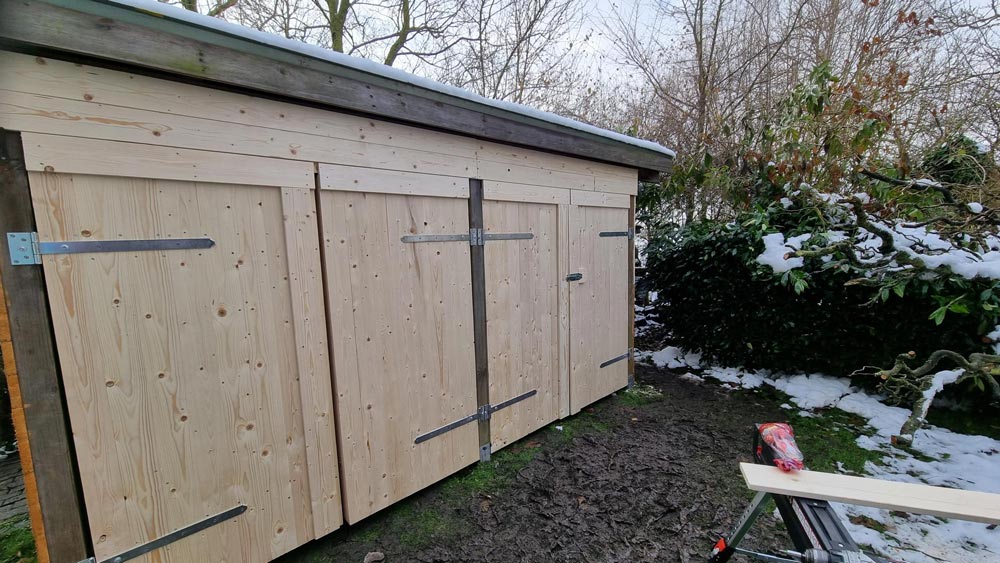 Eine hölzerne Gartenhütte mit neuen Türen, umgeben von Schnee und kahlen Bäumen.