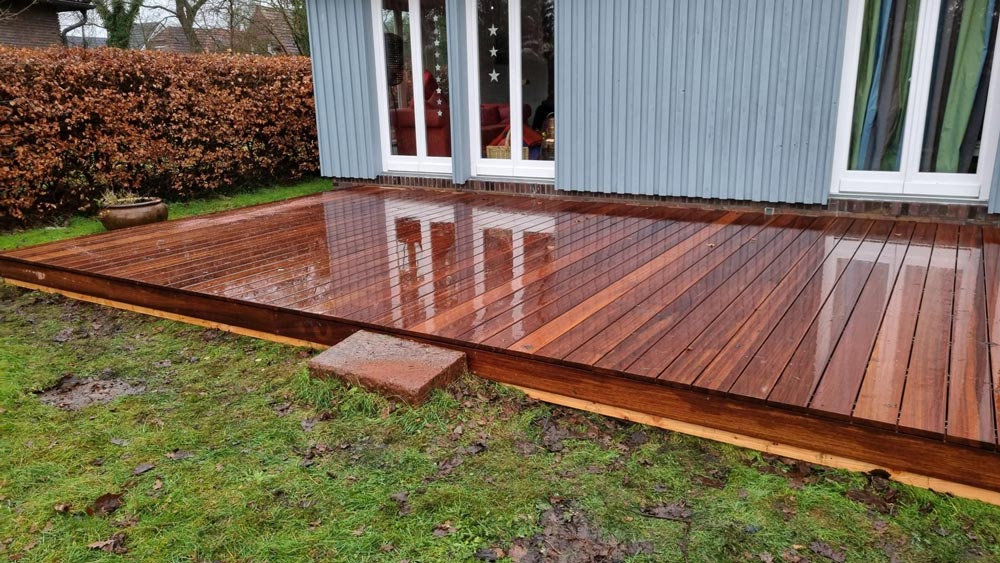 Nasses Holzdeck vor blauem Haus mit Hecken im Hintergrund. Feuchte, glänzende Oberfläche.